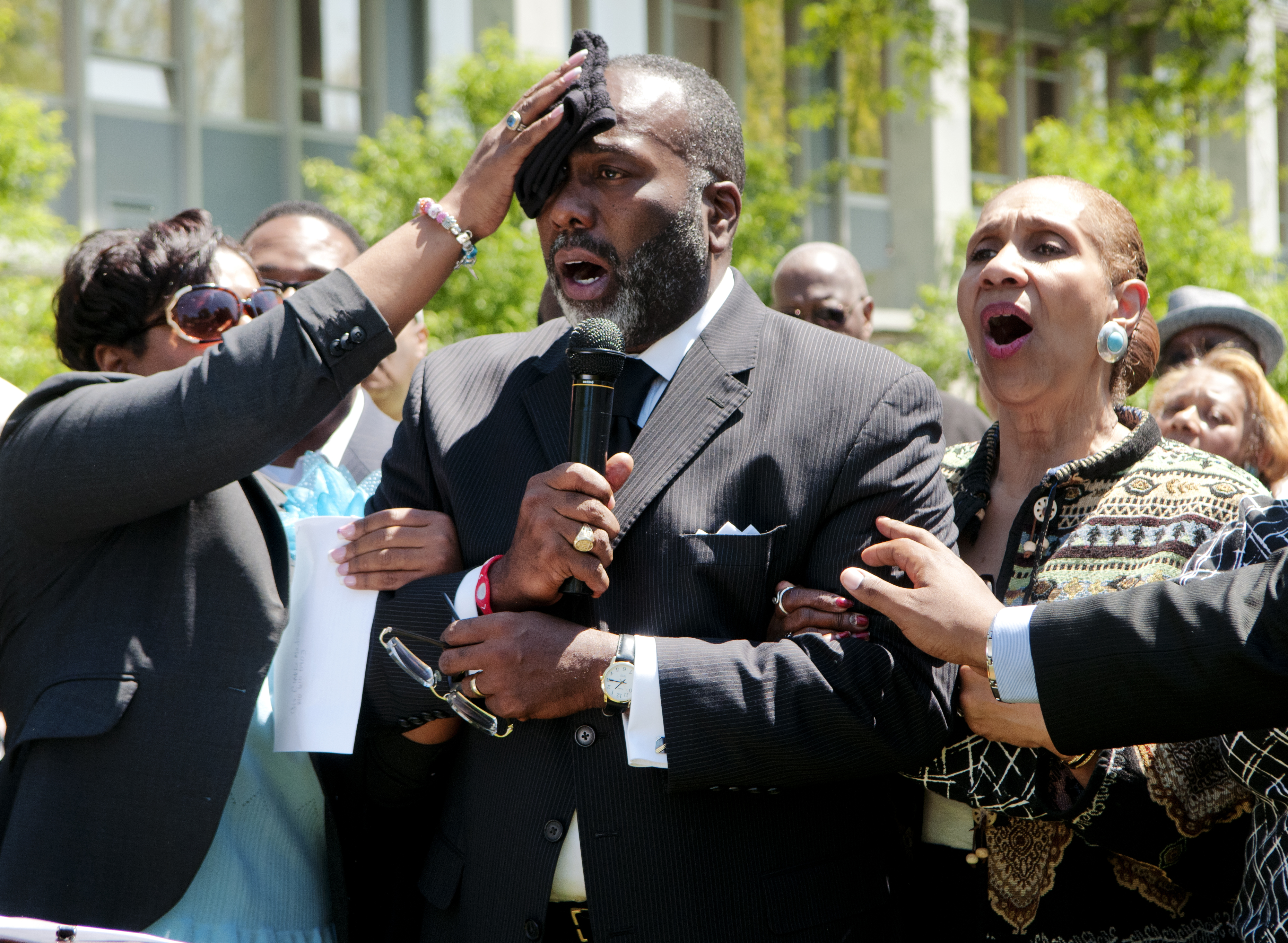 Delores Flynn wipes the forehead of her husband, Rev. Reginald Flynn, as he leads a crowd gathered on the front lawn of City Hall in Flint, Mich., in singing “We Shall Overcome” at the end of a rally against Public Act 4, May 14, 2012. (Ryan Garza/Flint Journal via AP)