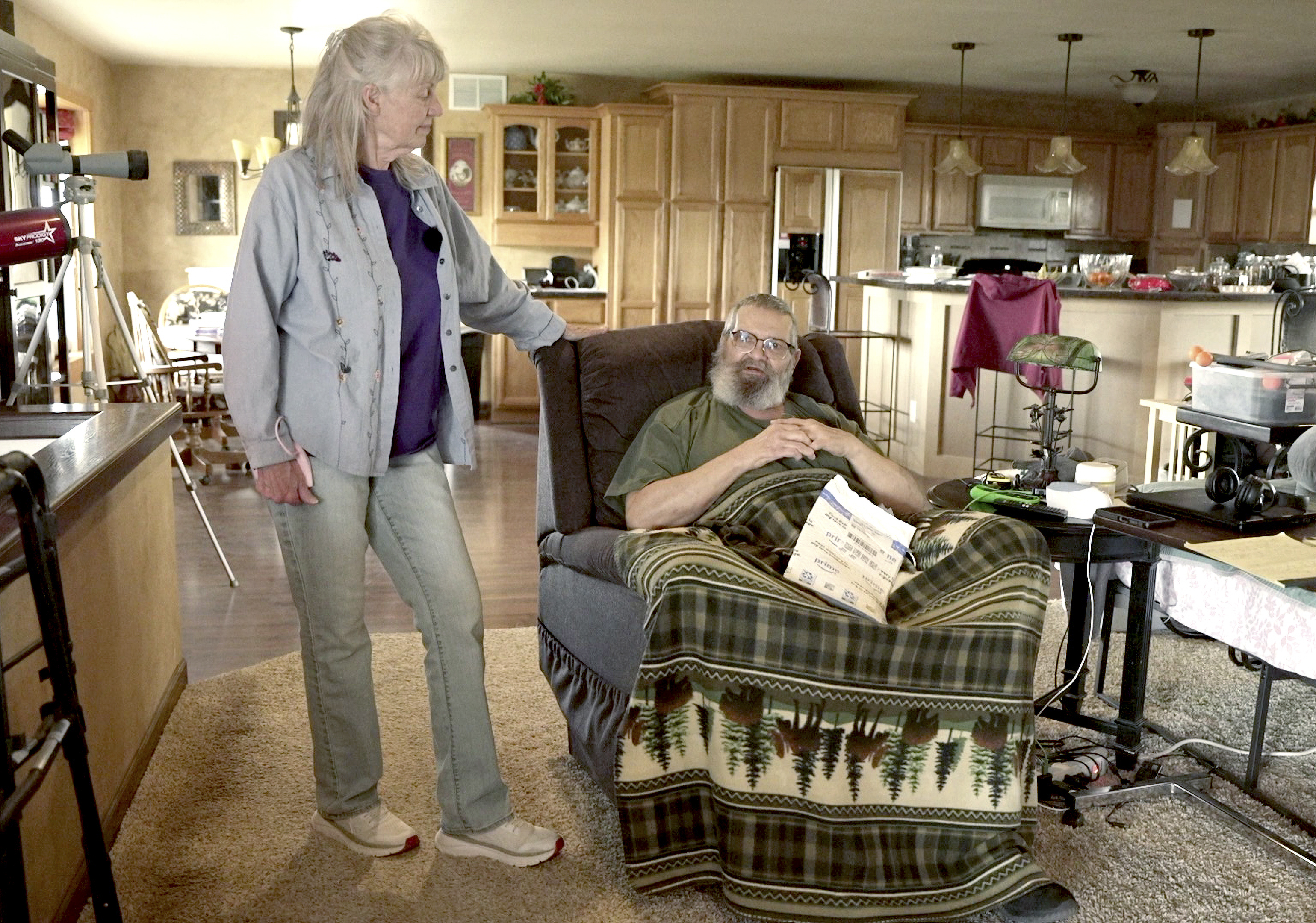 Gwen Smith (left) listens to her husband, Alan Smith, talk about their life in northwestern Nebraska after she walked a quarter mile to get the mail and gave him a small parcel on May 8, 2025. “It’s beautiful,” he said of the countryside where they live. The couple doesn’t get to town much due to Alan Smith’s health issues. They rely heavily on the Postal Service, especially for his medications.<br />(Nick Loomis / The Midwest Newsroom)