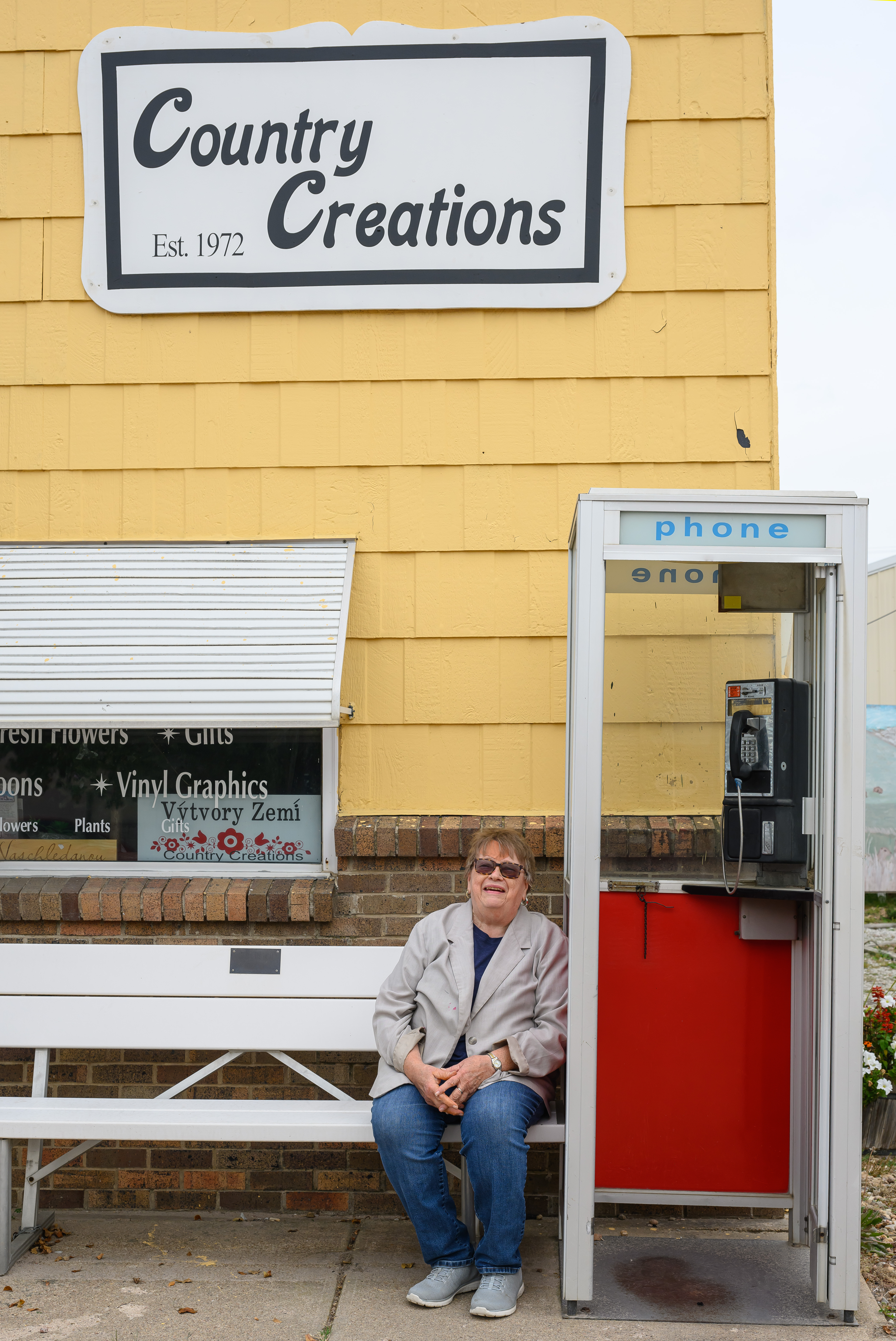 Delores Ruzicka, owner of the Country Creations store in downtown Verdigre, sits on a bench next to the non-working pay phone that stands outside her business on the town’s main street. Ruzicka said she often places change in the return slot for fun — and the coins always are gone the next time she checks.<br />(Darin Epperly / Flatwater Free Press)