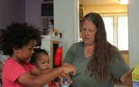 Liz Arnold interacts with two children (names withheld for privacy) at Just Like Mine Daycare in Lincoln, Nebraska, on July 22, 2026. She’s certified to care for up to 10 children in her home, but this summer she is taking care of six. 
(Daniel Wheaton / The Midwest Newsroom)
